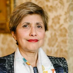 Portret kobiety z krótkimi brązowymi włosami, ubranej w ciemną marynarkę w pepitkę i białą apaszkę w kolorowe wzory, na tle ściany w kwiatowe wzory.