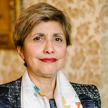 Portret kobiety z krótkimi brązowymi włosami, ubranej w ciemną marynarkę w pepitkę i białą apaszkę w kolorowe wzory, na tle ściany w kwiatowe wzory.