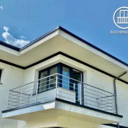 Nowoczesny balkon z metalową, minimalistyczną balustradą, biała elewacja budynku z czarnymi akcentami, błękitne niebo w tle.