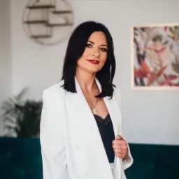 Kobieta w białym żakiecie, czarnej bluzce z koronką i złotym naszyjnikiem, z ciemnymi włosami i czerwoną szminką, pozująca na tle jasnej ściany z abstrakcyjnym obrazem i zieloną sofą.