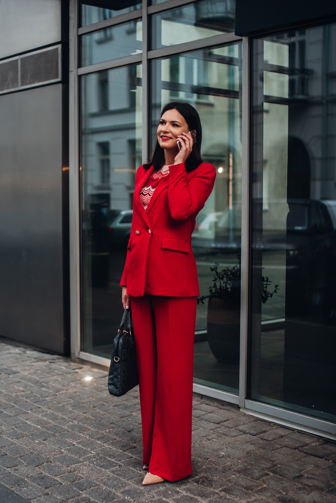 Uśmiechnięta kobieta w czerwonym garniturze rozmawia przez telefon na tle nowoczesnego budynku z szklaną fasadą. W ręku trzyma czarną torebkę.