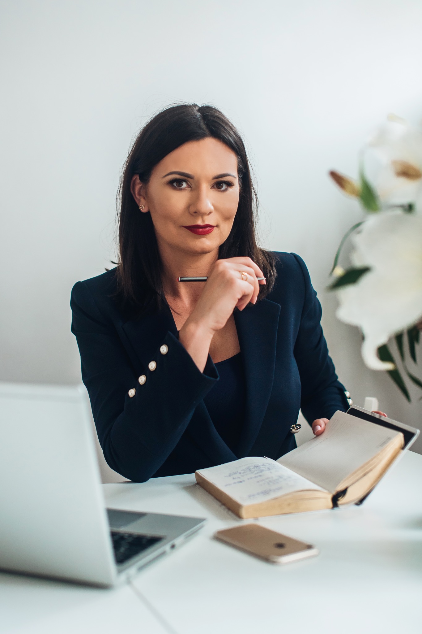 Kobieta w granatowej marynarce siedzi przy biurku z laptopem i notatnikiem, trzymając długopis przy ustach. W tle białe kwiaty i jasne tło.