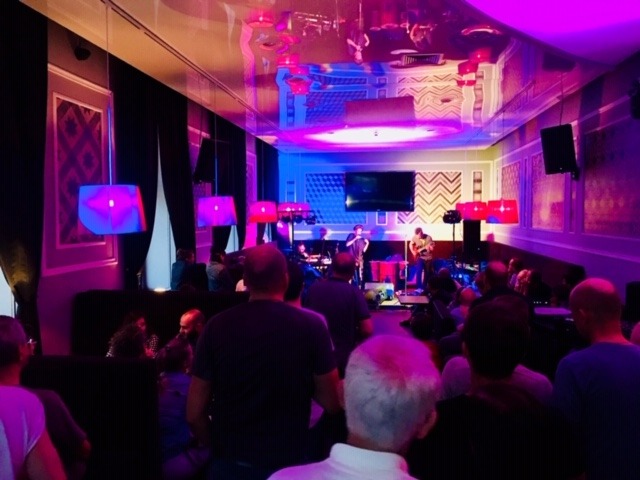 Występ zespołu muzycznego na żywo podczas wydarzenia firmowego w Łodzi, oświetlenie sceniczne w odcieniach fioletu i różu, widownia siedząca przy stolikach.