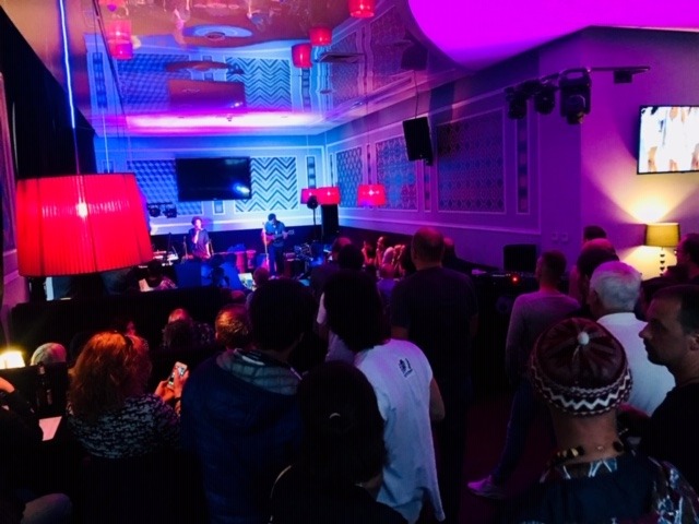 Zespół muzyczny na scenie oświetlonej czerwonymi i niebieskimi światłami, grający dla publiczności w eleganckim wnętrzu z wzorzystymi ścianami.