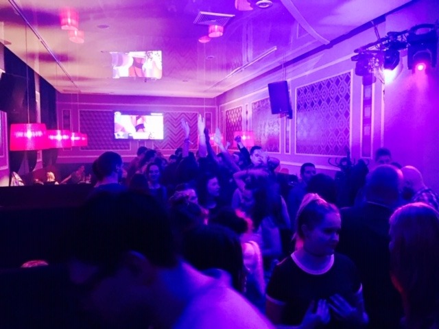 Wnętrze klubu nocnego wypełnione tańczącymi ludźmi, oświetlone intensywnym fioletowym światłem, z ekranami wyświetlającymi obrazy na ścianach i ozdobnymi lampami wiszącymi z sufitu.