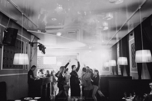 Czarno-białe zdjęcie z imprezy w klubie, widoczni ludzie tańczący w dymie i świetle reflektorów, DJ przy konsolecie po lewej stronie, dekoracyjne lampy wiszące z sufitu.