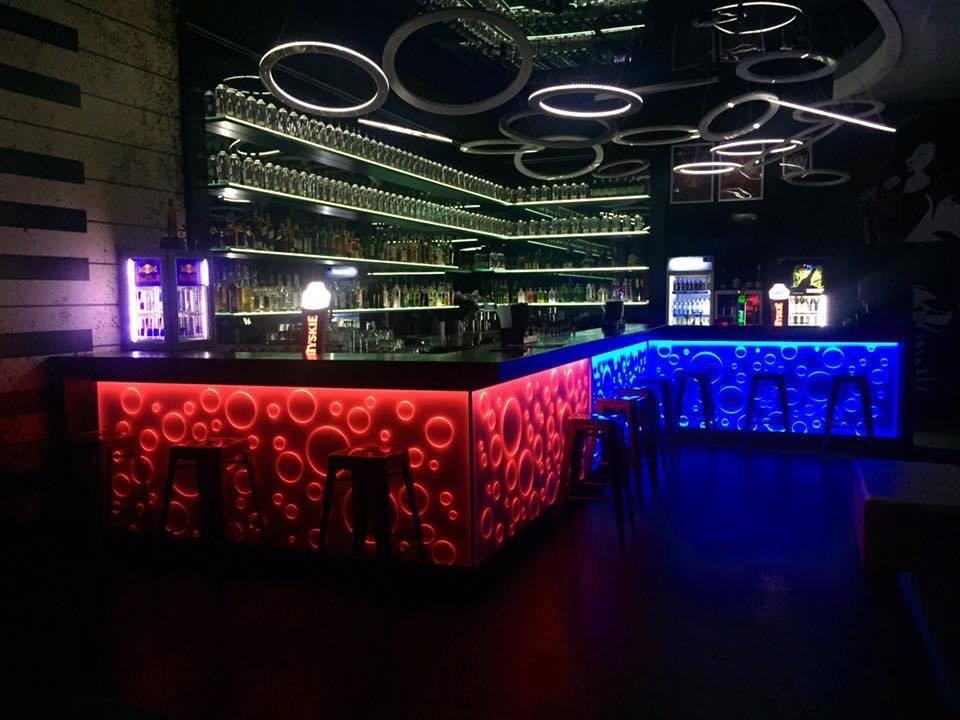 Podświetlony bar z okrągłymi wzorami, czerwony z lewej i niebieski z prawej strony, półki z butelkami w tle, okrągłe lampy na suficie.