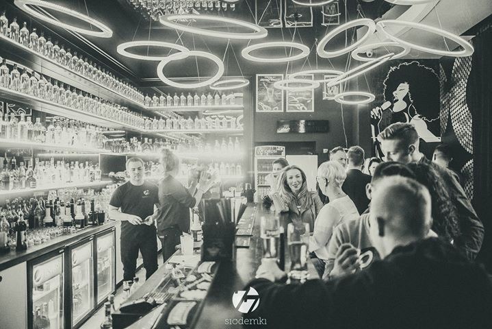 Czarno-białe zdjęcie wnętrza baru z klientami przy barze, podświetlonymi półkami z butelkami i dekoracyjnymi lampami w kształcie okręgów
