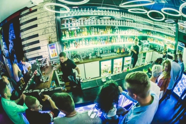 Wnętrze baru z wystrojem w stylu fortepianu, oświetlone neonowymi lampami, barman w czarnej koszulce przygotowuje drinki dla klientów zgromadzonych przy barze.