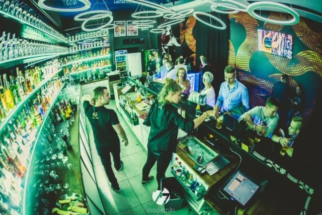 Wnętrze baru z oświetleniem neonowym, obsługa przygotowuje drinki dla gości siedzących przy ladzie, na półkach widoczne liczne butelki z alkoholem.