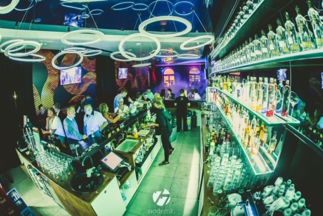 Wnętrze baru z oświetleniem neonowym, pełne półki z butelkami alkoholu, obsługa za barem i klienci siedzący przy ladzie.