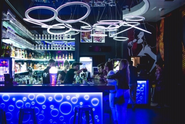 Wnętrze baru z podświetlanym na niebiesko blatem, barman nalewa drinki, w tle półki z alkoholem i dekoracyjne lampy w kształcie okręgów.