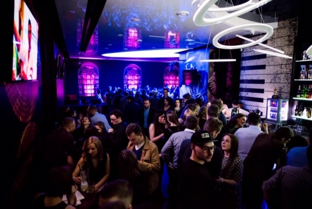 Wnętrze klubu nocnego z dużą ilością osób, podświetlonym barem i dekoracyjnymi lampami pierścieniowymi na suficie.