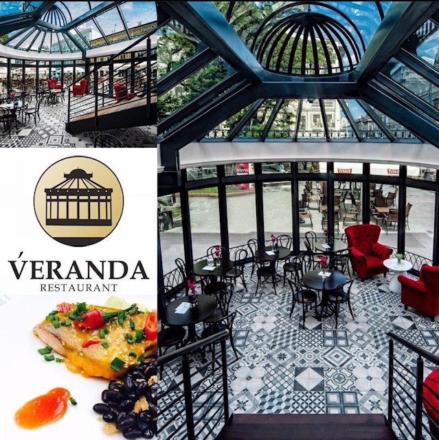 Wnętrze restauracji Veranda z przeszklonym dachem, czarnymi stolikami i krzesłami, wzorzystą podłogą oraz logo restauracji i zdjęciem dania z ryby.