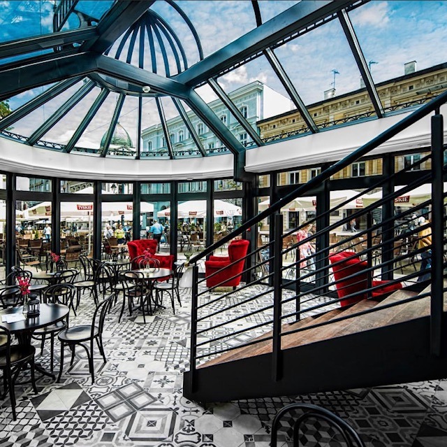 Wnętrze kawiarni z przeszklonym dachem, wzorzystą posadzką, czarnymi stolikami i krzesłami oraz czerwonymi fotelami, widok na ulicę przez okna.