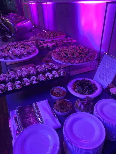 Bufet z przekąskami na stole podświetlonym fioletowym światłem, widoczne talerze z różnorodnymi przystawkami, miseczki z dipami, stosy talerzy i sztućce.