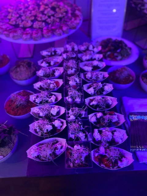 Stół z różnorodnymi przekąskami w małych naczyniach, w tym sałatki i tartaletki, oświetlony fioletowym światłem, tworząc elegancką prezentację.