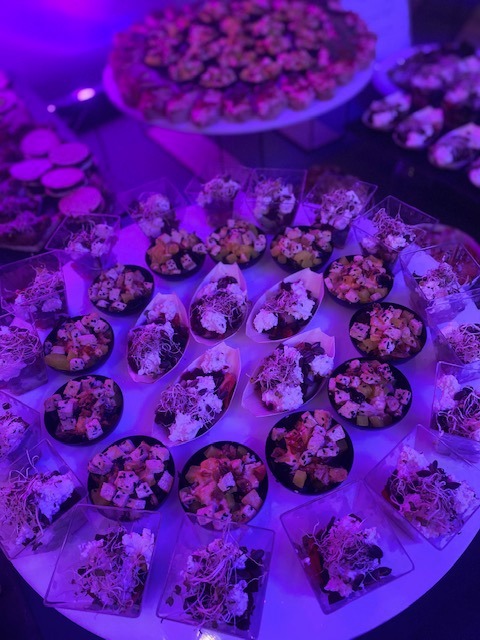 Stół z różnorodnymi przekąskami w małych naczyniach, oświetlony fioletowym światłem, widoczne sałatki i tartinki w różnych kształtach i kolorach.