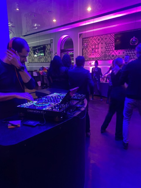 DJ z pomarańczowymi słuchawkami obsługuje konsoletę Denon DJ na tle tańczących ludzi w lokalu z fioletowym oświetleniem.