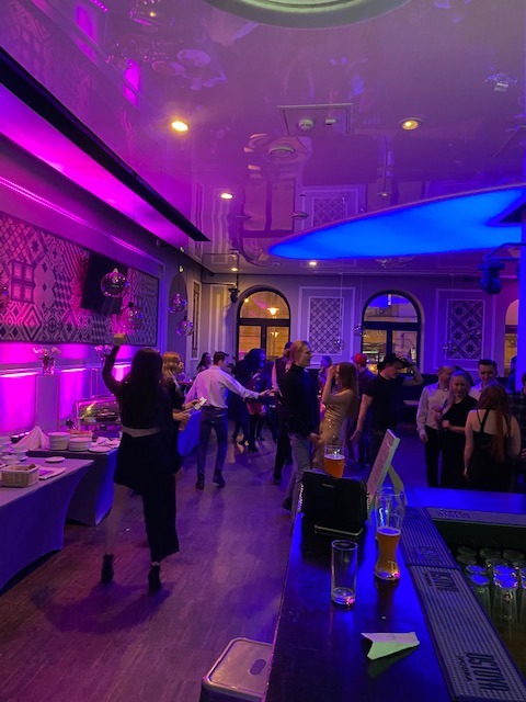 Tłum ludzi tańczących w lokalu oświetlonym fioletowymi i niebieskimi światłami, widoczny bar z napojami i catering w tle.