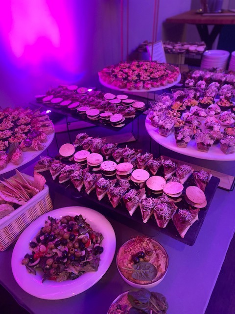 Stół z różnorodnymi przekąskami: sałatka z oliwkami, hummus z dekoracją z liści, mini kanapki z okrągłymi ciastkami, przekąski w szklanych pojemnikach, oświetlony fioletowym światłem.