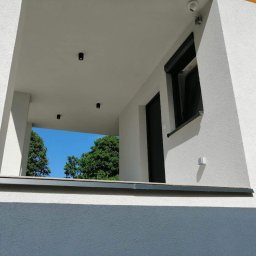 Tom-Kos - Nowoczesna elewacja domu z widocznym fragmentem balkonu, czarnymi drzwiami, oknem z roletą zewnętrzną i punktowym oświetleniem zewnętrznym.