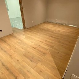 Tom-Kos - Jasne drewniane panele podłogowe ułożone w nowym, jeszcze nieumeblowanym pokoju z pomalowanymi ścianami w odcieniach beżu i szarości.