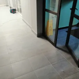 Szare płytki podłogowe ułożone w korytarzu, widoczny kabel zasilający i urządzenie czyszczące w tle, otwarte drzwi z tabliczką ostrzegawczą 'Uwaga ślisko'.