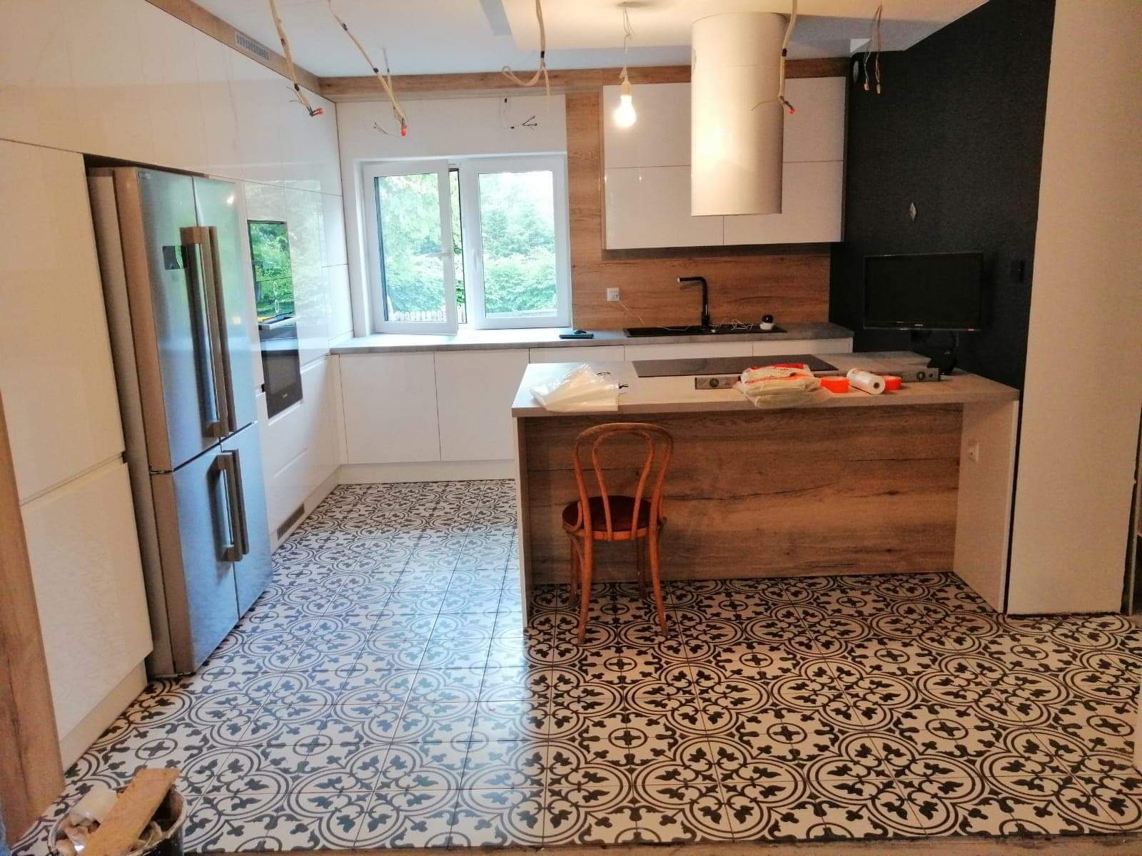 Nowoczesna kuchnia w trakcie wykańczania: białe szafki, drewniane akcenty, wyspa z blatem i wzorzysta podłoga w stylu retro, widoczne kable i lampa bez klosza.