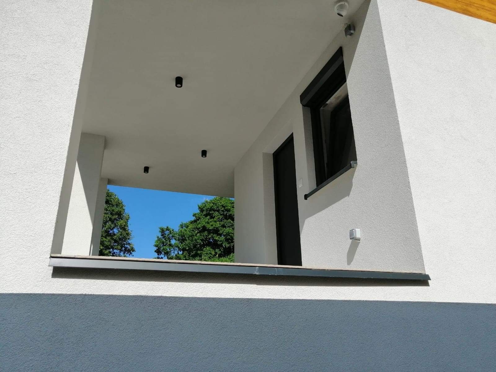 Nowoczesna elewacja domu z widocznym fragmentem balkonu, czarnymi drzwiami, oknem z roletą zewnętrzną i punktowym oświetleniem zewnętrznym.