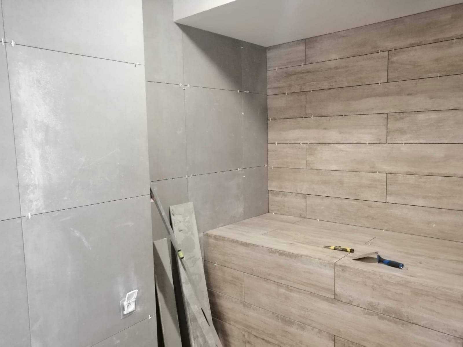 Łazienka w trakcie wykańczania: szare, prostokątne płytki na ścianie i obudowie wnęki, krzyżyki dystansowe widoczne w fugach, narzędzia glazurnicze leżą na obudowie.