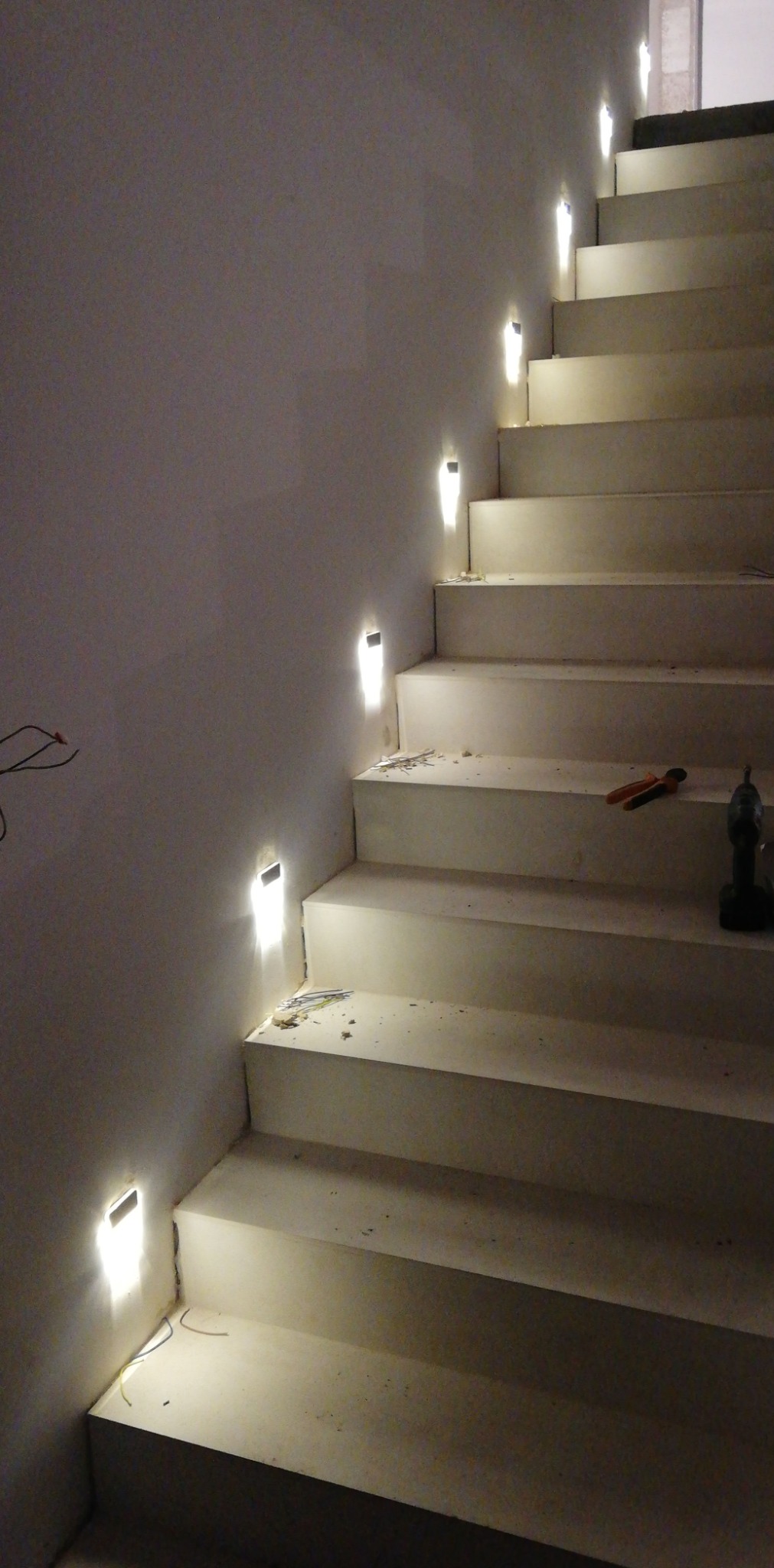 Schody z zamontowanym oświetleniem LED w ścianie, widoczne narzędzia: wiertarka i kombinerki, przewody elektryczne wystające ze ściany.
