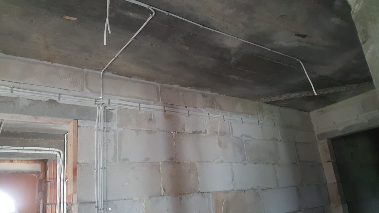 Surowe wnętrze w trakcie prac: widoczne białe przewody elektryczne poprowadzone po ścianach z bloczków i betonowym suficie, przygotowane do dalszej instalacji.
