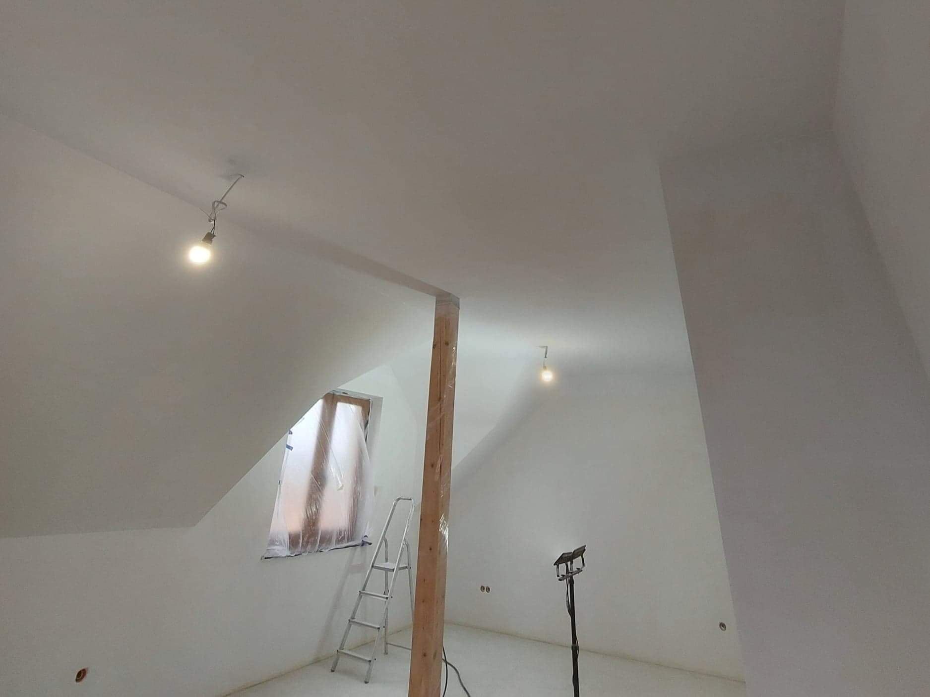 Poddasze po zagruntowaniu ścian, z zabezpieczonym oknem folią malarską, drabina oparta o ścianę, widoczna drewniana kolumna i lampa halogenowa na statywie.