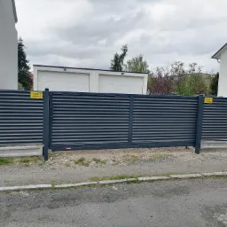 Przesuwna brama z poziomych, szarych paneli, z żółtymi tabliczkami ostrzegawczymi, zamontowana na betonowej podmurówce, widok z zewnątrz na tle zabudowań.