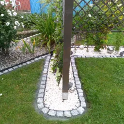 Obrzeże z kostki granitowej oddzielające trawnik od rabaty z białym grysem, w tle drewniana pergola z pnącymi roślinami i fragment ogrodzenia panelowego.