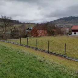 Długi, czarny płot z siatki na betonowej podmurówce, biegnący wzdłuż zbocza wzgórza, z widokiem na domy i zalesione wzgórza w tle, pochmurny dzień.