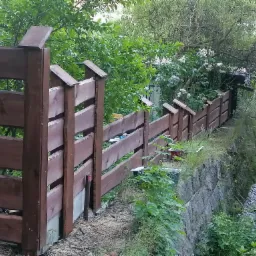 Nowo postawione, brązowe ogrodzenie z drewnianych desek na betonowej podmurówce, biegnące wzdłuż kamiennego muru i strumienia, otoczone bujną zielenią.