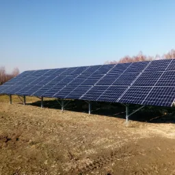 Instalacja fotowoltaiczna na gruncie z widocznymi panelami słonecznymi, stalową konstrukcją wsporczą i częściowo zoranym podłożem w słoneczny dzień.
