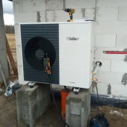 Biała pompa ciepła Vaillant aroTHERM zamontowana na betonowych bloczkach, z podłączonym miernikiem parametrów i wiertarką DeWalt na górze, na tle ściany z bloczków betonowych i krajobrazu.