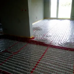 Rozłożone rury ogrzewania podłogowego na siatce w surowym pomieszczeniu z oknem, zabezpieczone folią, widoczne czerwone rury wzdłuż ścian.