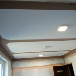 Kry-Bud - Wnętrze pokoju w trakcie prac wykończeniowych, widoczne pomalowane listwy sufitowe w kolorze brązowym, sufit z oświetleniem LED, ściany przygotowane do malowania, oraz częściowo widoczne drzwi.