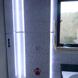 Kry-Bud - Łazienka w trakcie wykańczania z podświetlanymi pionowymi pasami płytek, widoczne okno z zabezpieczeniem folią, instalacja sanitarna i fragment wanny.