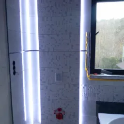 Łazienka w trakcie wykańczania z podświetlanymi pionowymi pasami płytek, widoczne okno z zabezpieczeniem folią, instalacja sanitarna i fragment wanny.