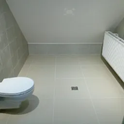 Wykończona łazienka na poddaszu ze ścianami i podłogą wyłożonymi szarymi płytkami, wisząca toaleta i grzejnik.