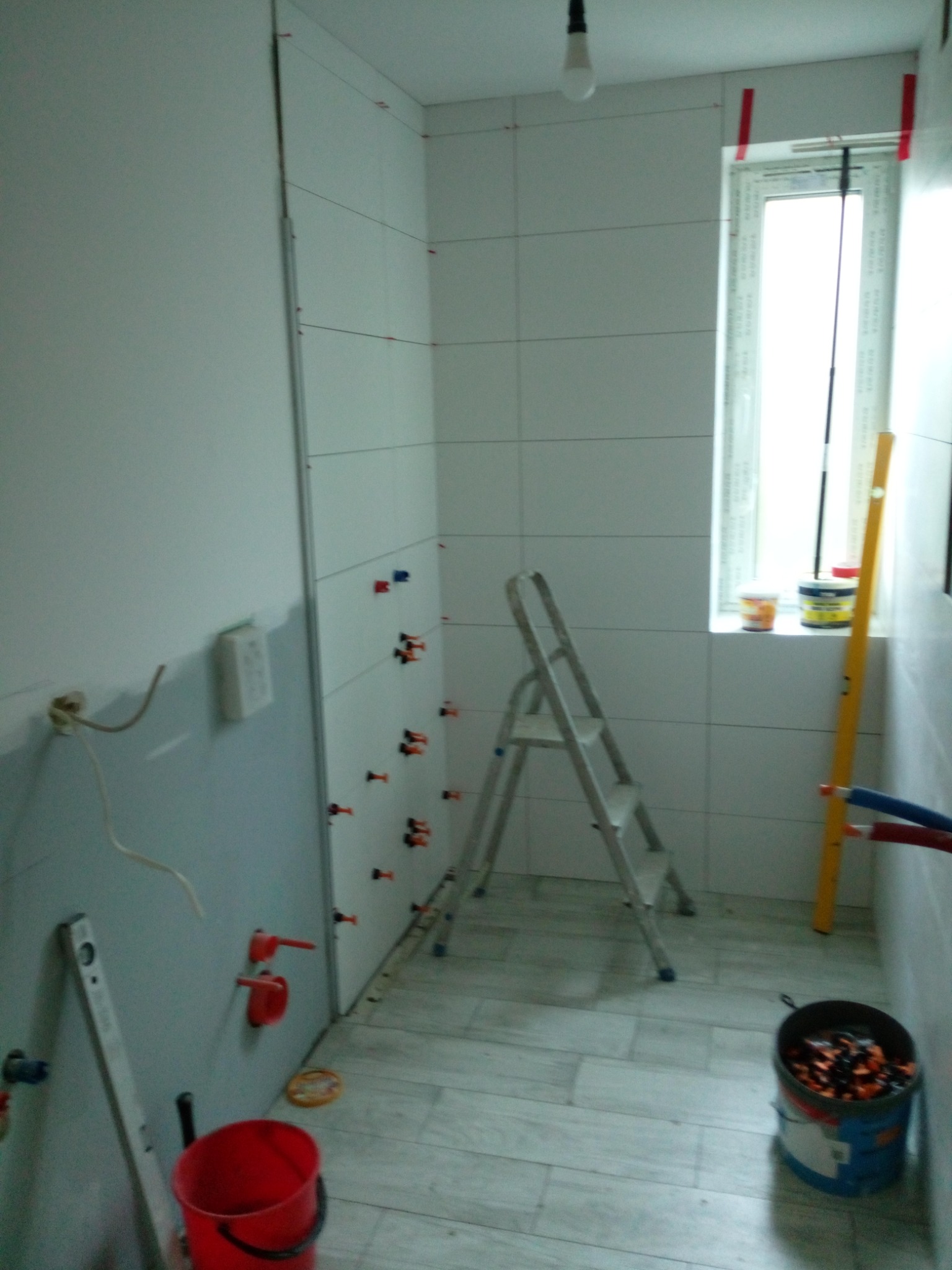 Łazienka w trakcie układania białych płytek z użyciem krzyżyków dystansowych, widoczna drabina, poziomica, wiadro i puszki z materiałami budowlanymi.