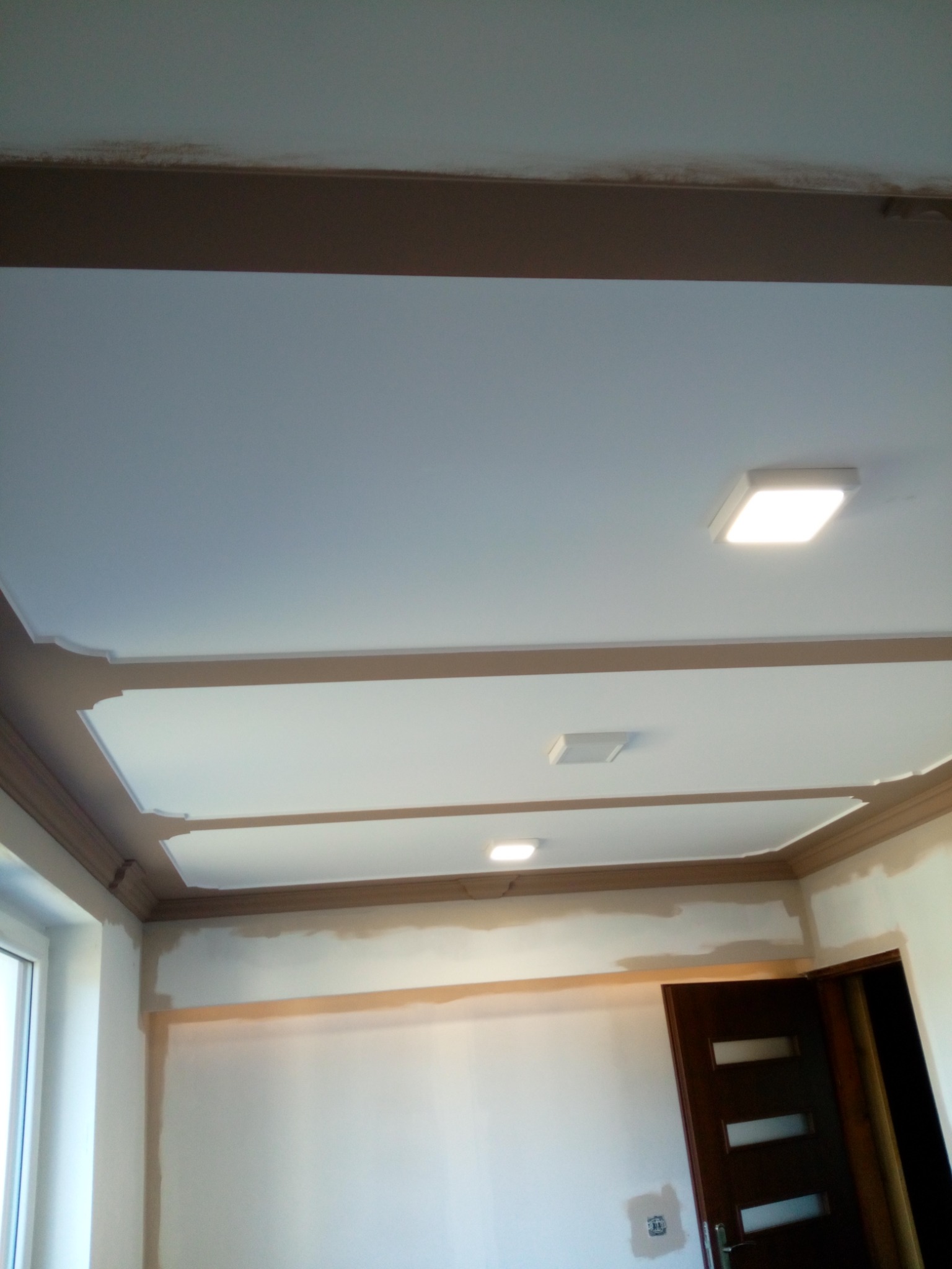 Wnętrze pokoju w trakcie prac wykończeniowych, widoczne pomalowane listwy sufitowe w kolorze brązowym, sufit z oświetleniem LED, ściany przygotowane do malowania, oraz częściowo widoczne drzwi.
