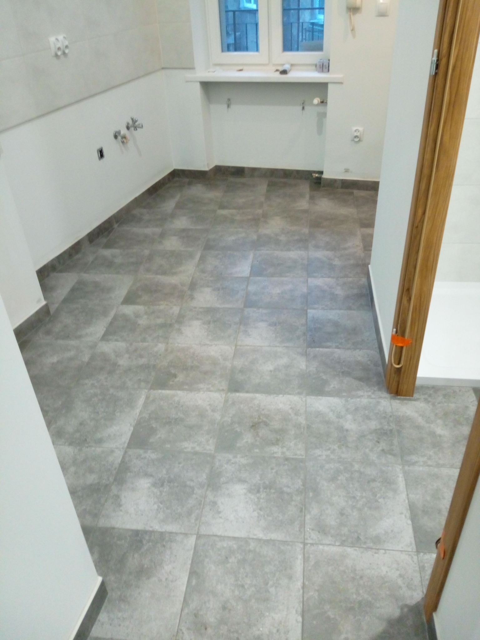 Szare, kwadratowe płytki podłogowe w łazience, widoczne instalacje wodno-kanalizacyjne na ścianie i fragment drewnianej ościeżnicy drzwiowej z pomarańczowym zabezpieczeniem.