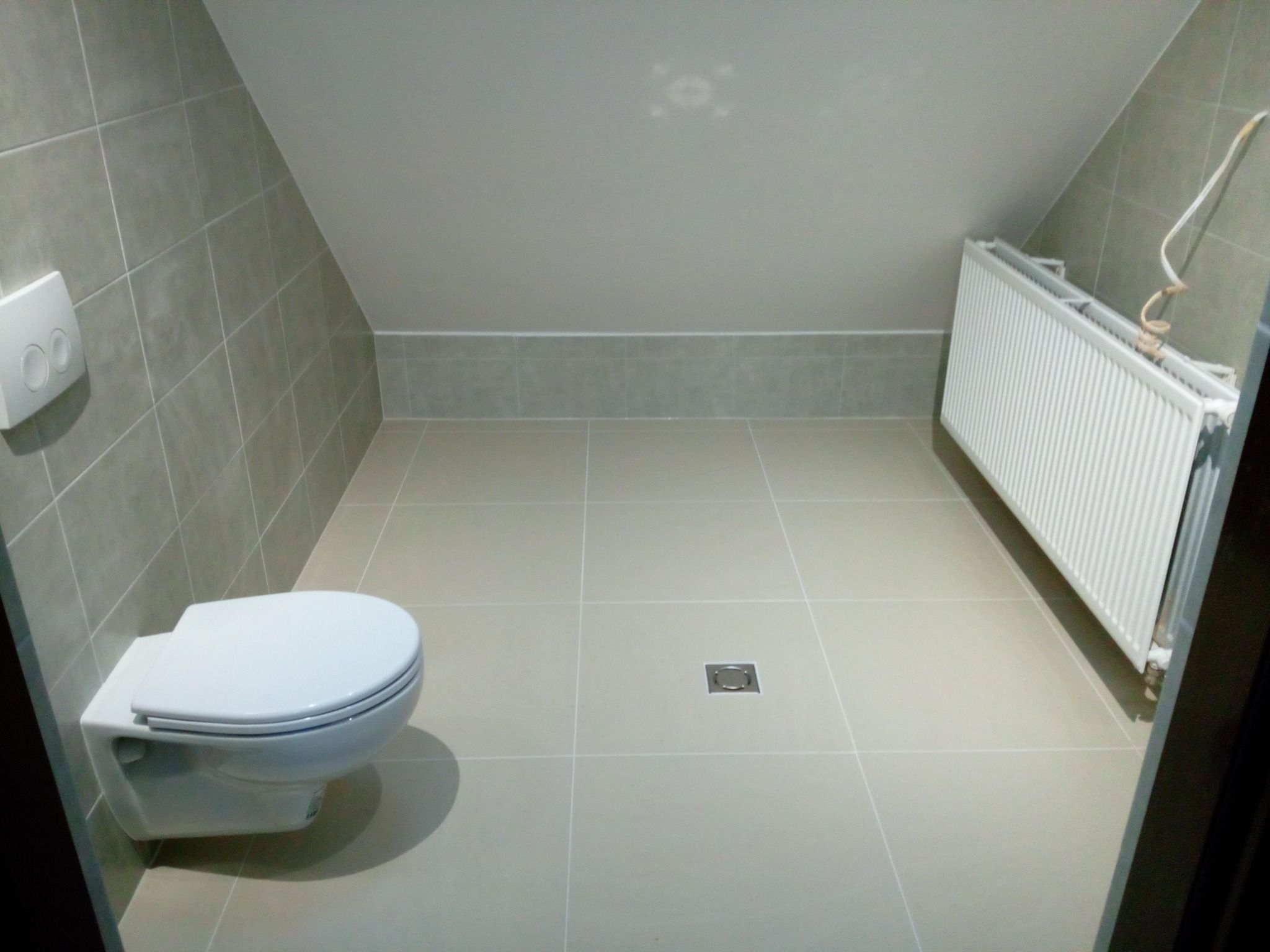 Wykończona łazienka na poddaszu ze ścianami i podłogą wyłożonymi szarymi płytkami, wisząca toaleta i grzejnik.