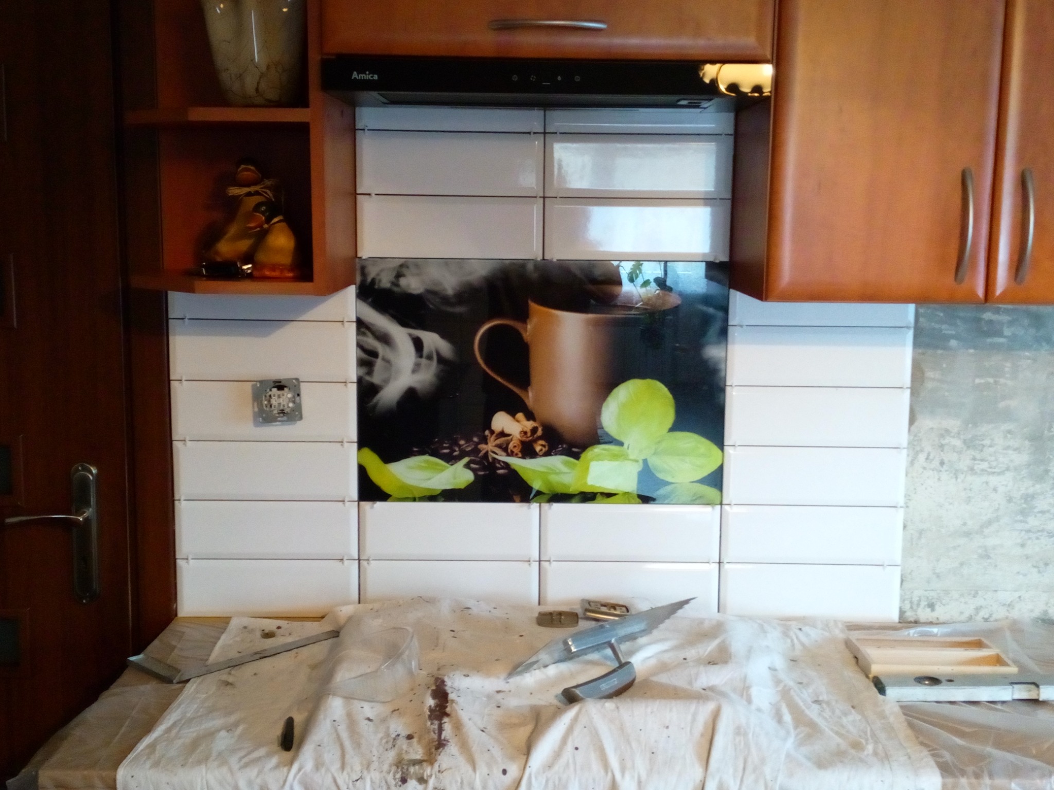 Układanie białych płytek w kuchni w Pcimiu, z dekoracyjnym panelem z motywem kawy i narzędziami glazurniczymi na blacie roboczym.